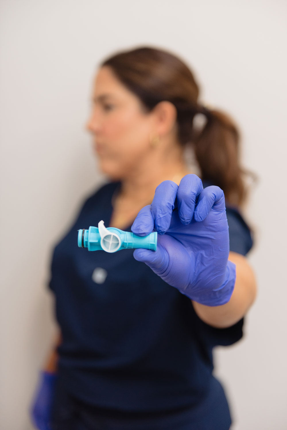 A person wearing blue medical gloves holds a small light blue and white dental device toward the camera, with a blurred clinical background and a person in navy scrubs out of focus behind them.