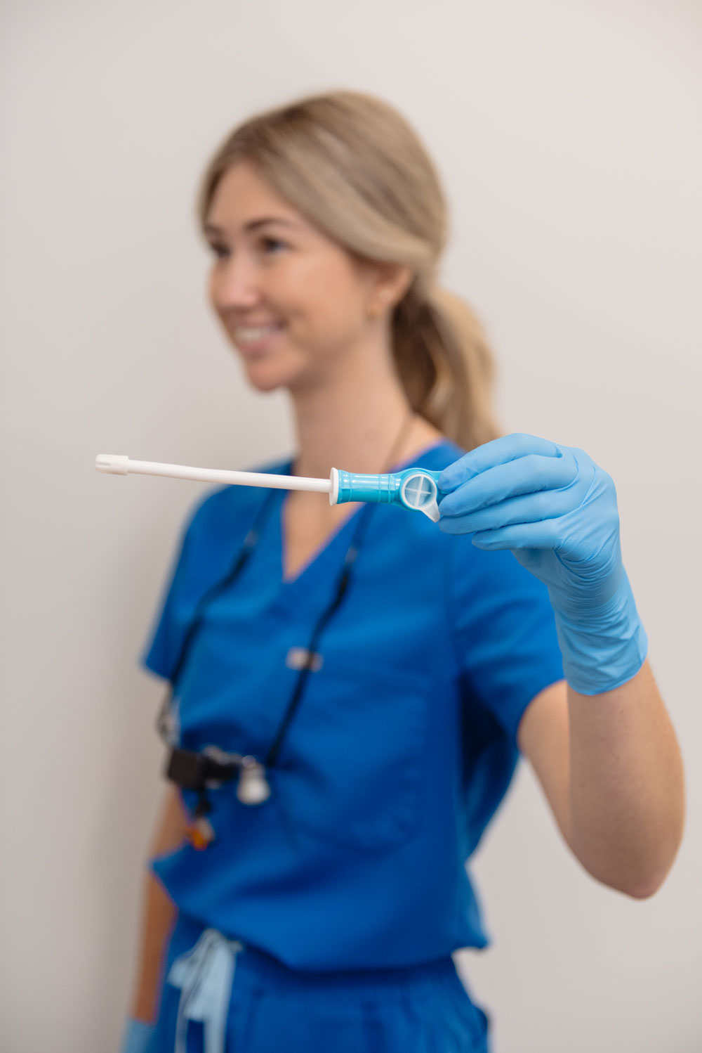 A healthcare worker in blue scrubs holds a blue and white dental device with a white handle, wearing blue gloves against a plain light gray background.