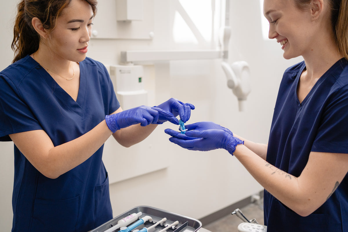 Two healthcare workers in navy blue scrubs and purple gloves handling a small dental valve in a bright clinical room. #Color_Blue