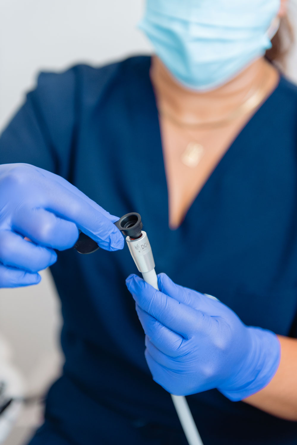 A close-up of a person wearing blue medical scrubs, a light blue face mask, and blue gloves, holding and demonstrating a tailcap cover.
