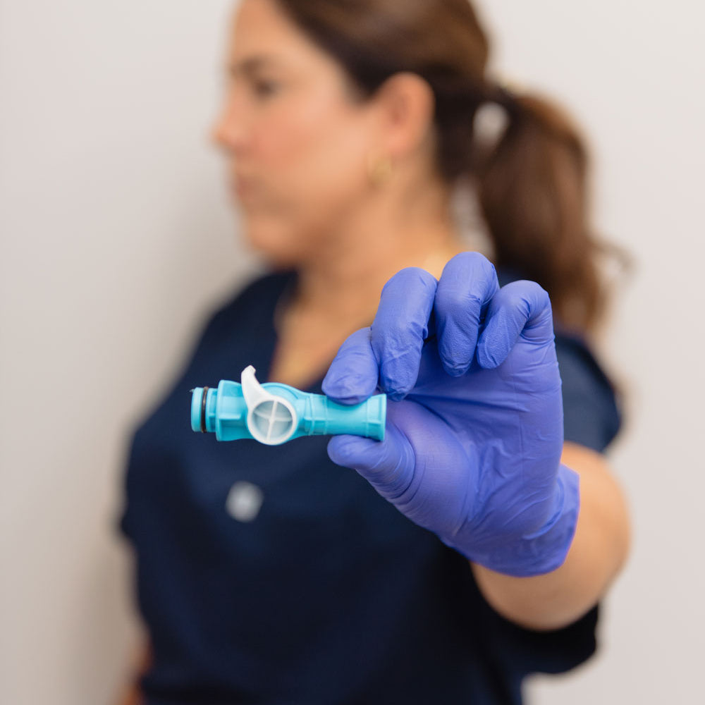 A person wearing blue medical gloves holds a small light blue and white dental device toward the camera, with a blurred clinical background and a person in navy scrubs out of focus behind them.