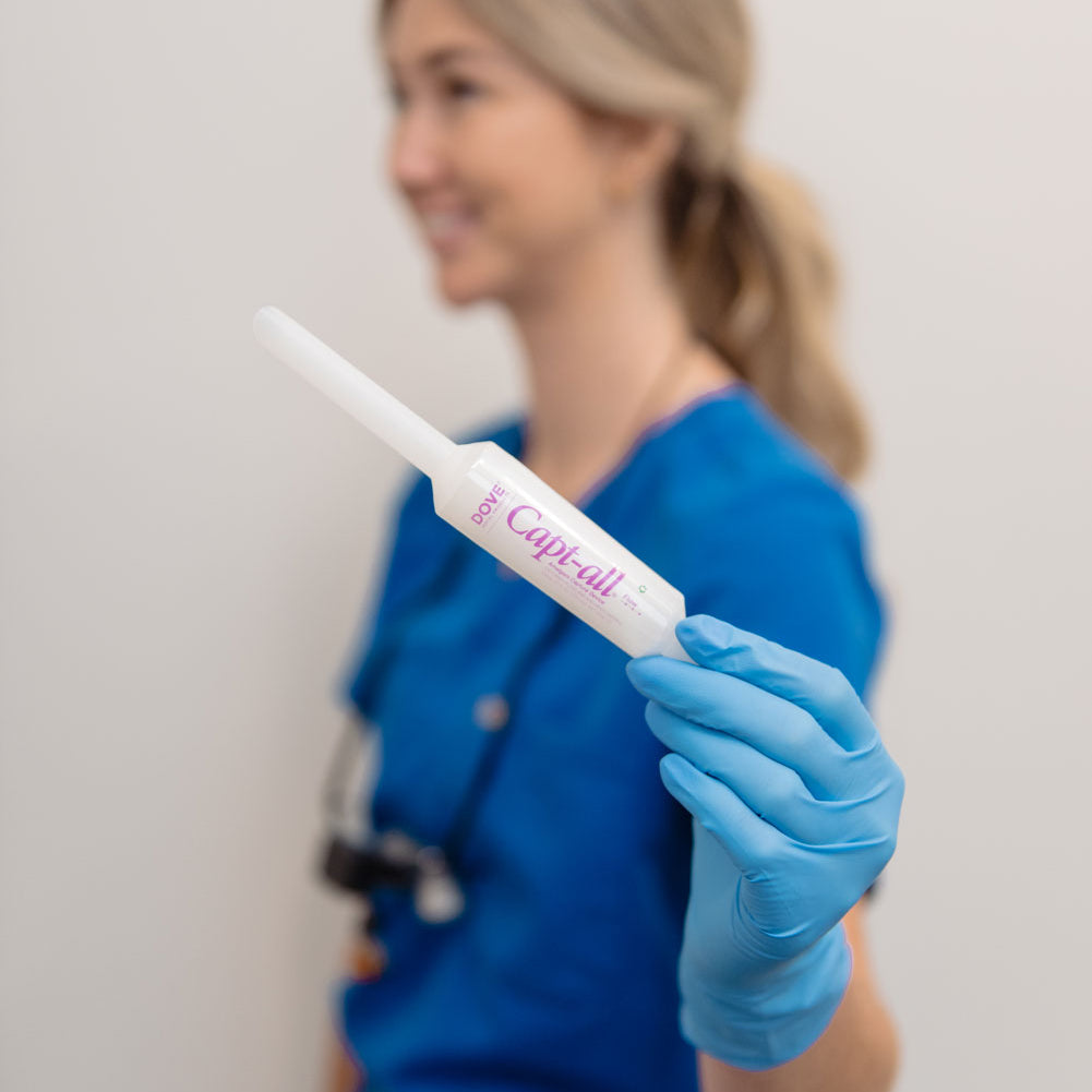 A woman in blue medical scrubs and a blue glove holds a Capt-all dental device, photographed against a plain light background with the subject softly blurred.