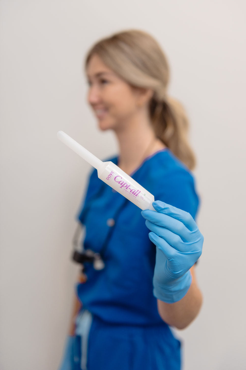 A woman in blue medical scrubs and a blue glove holds a Capt-all dental device, photographed against a plain light background with the subject softly blurred.